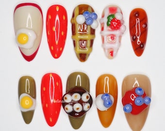 Peut inclure: Dix faux ongles avec des motifs sur le thème de la nourriture. Les motifs comprennent un œuf au plat, des fraises, des gaufres aux myrtilles et une sauce rouge. Les ongles sont de différentes couleurs, dont le rouge, le marron et le beige.