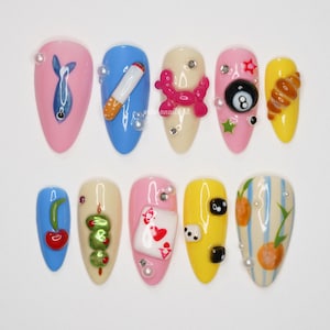 May include: A collection of ten almond-shaped artificial nails, each in a different colour and decorated with unique 3D designs. Designs include a fish, a cigarette, a balloon dog, an 8-ball, a croissant, a cherry, olives, a playing card, dice, and oranges.