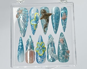 May include: A set of ten long, pointed artificial nails in a clear plastic case. The nails feature a variety of ocean-themed designs, including blue and white swirls, an octopus, and seashell embellishments. Some nails have 3D elements.