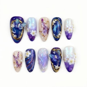 May include: A set of ten artificial nails with various designs. The nails feature butterfly, floral, and abstract patterns in shades of blue, purple, and white. Some nails have gold accents and pearl embellishments.
