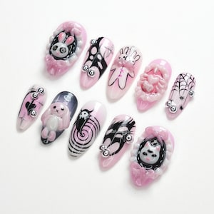 May include: A collection of ten artificial nails with various designs. The nails are primarily pink and feature black and white illustrations of rabbits, spiders, and other gothic-themed elements. Some nails have 3D embellishments.