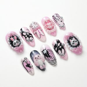 May include: A collection of pink and white artificial nails with various designs. Some nails feature 3D elements like a bunny, spiderweb, and heart. Other nails have black and white graphic designs and silver embellishments. The nails are arranged on a white surface.