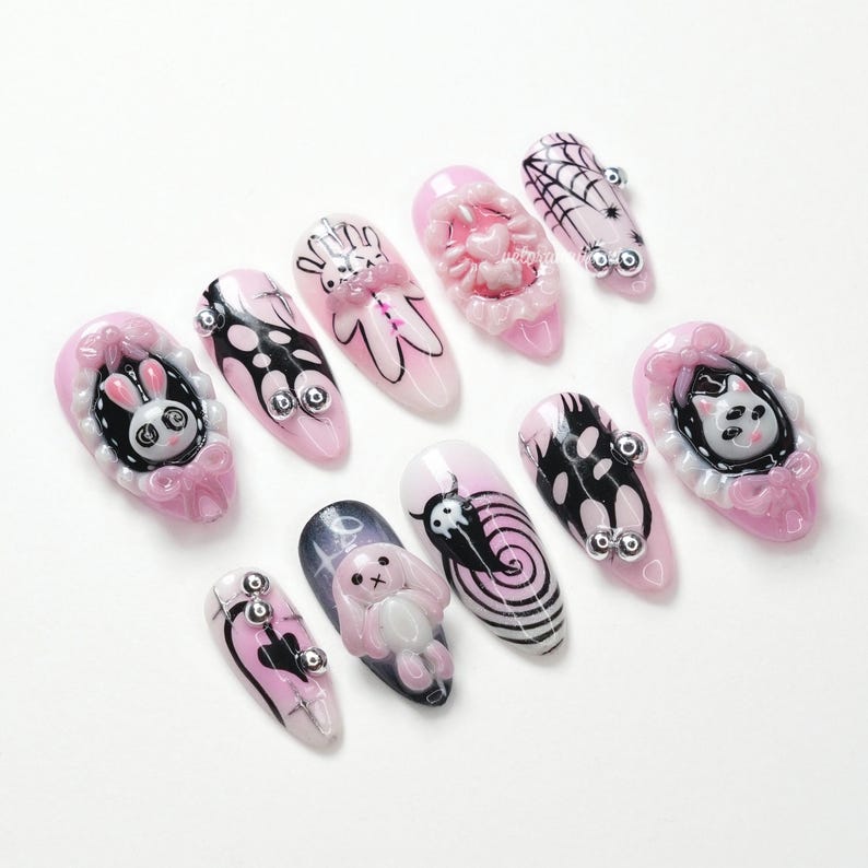 May include: A collection of pink and white artificial nails with various designs. Some nails feature 3D elements like a bunny, spiderweb, and heart. Other nails have black and white graphic designs and silver embellishments. The nails are arranged on a white surface.