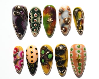 Op de afbeelding: Een verzameling van tien amandelvormige, beschilderde vingernagels. Elke nagel heeft unieke ontwerpen met verschillende kleuren, waaronder paars, groen, geel en oranje. Sommige nagels hebben gouden accenten en versieringen, terwijl andere ingewikkelde patronen en texturen vertonen.