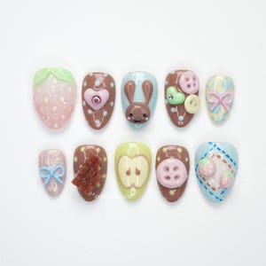 May include: A collection of ten press-on nails with diverse designs. The nails showcase a strawberry, a heart, a bunny, buttons, bows, chocolate, an apple, and strawberries. Colours include pink, brown, blue, yellow, and green. The nails are displayed on a white surface.