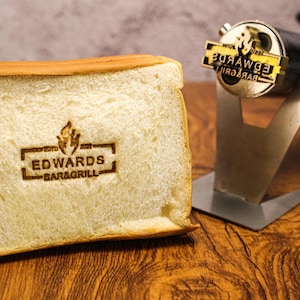 Könnte beinhalten: Eine Brotscheibe mit dem Logo "EDWARDS BAR & GRILL" eingebrannt. Das Brot ist hellbraun mit goldener Kruste. Ein Metall-Brandeisen befindet sich im Hintergrund.