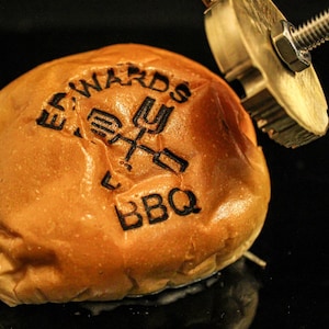 May include: A close-up shot of a golden-brown hamburger bun with the words "EDWARDS BBQ" branded on top. A metal branding iron is positioned above the bun, ready to be used. The bun sits on a dark surface.