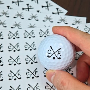 May include: A white golf ball with a dimpled surface, featuring the initials "C.F." and crossed golf clubs in black. Sheets of matching stickers with the same design are in the background. Golf-themed.
