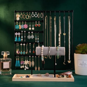 May include: A black metal jewelry organizer displaying various earrings and necklaces. The earrings are in different styles and colors, including gold hoops, floral designs, and tassel earrings. Necklaces include delicate chains and beaded strands. A wooden tray holds rings and other accessories.