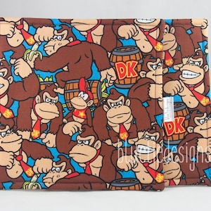 May include: A set of two brown and blue fabric coasters featuring a repeating pattern of Donkey Kong characters, barrels, and bananas. The coasters have a blue background with a white border.