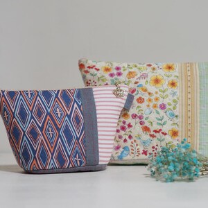 May include: Two fabric pouches with different patterns. One has a blue and orange diamond design, the other features a floral pattern with birds. A small bunch of blue dried flowers is in front of the pouches.