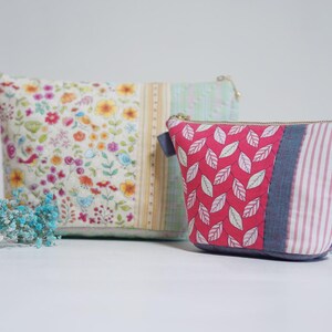 May include: Two quilted fabric pouches. The larger pouch features a floral pattern with birds, while the smaller one has a red leaf design. Both have zipper closures and are in shades of white, pink, and blue.