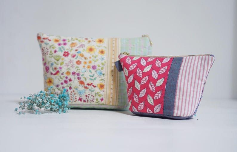 May include: Two quilted fabric pouches. The larger pouch features a floral pattern with birds, while the smaller one has a red leaf design. Both have zipper closures and are in shades of white, pink, and blue.