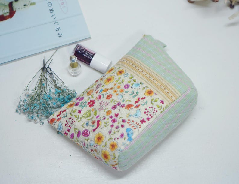 May include: A floral print cosmetic bag with a light green quilted exterior. The bag features a colorful pattern of flowers and birds. A bottle of nail polish and a small perfume bottle are next to the bag.
