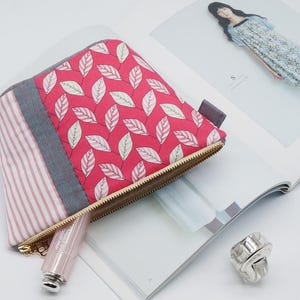 May include: A small, quilted cosmetic bag with a red leaf pattern, a gray stripe, and a pink and white striped section. The bag has a gold zipper and a small gray tab. A tube of lipstick and a silver ring are also visible.