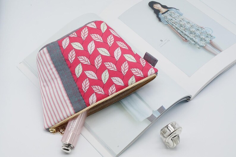 May include: A small, quilted cosmetic bag with a red leaf pattern, a gray stripe, and a pink and white striped section. The bag has a gold zipper and a small gray tab. A tube of lipstick and a silver ring are also visible.