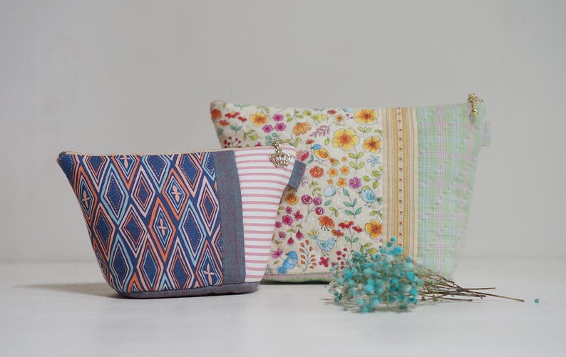 May include: Two fabric pouches with different patterns. One has a blue and orange diamond design, the other features a floral pattern with birds. A small bunch of blue dried flowers is in front of the pouches.