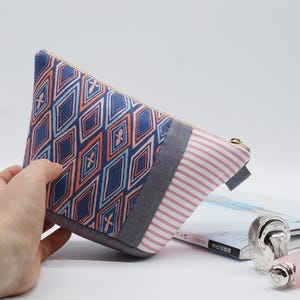 May include: A triangular cosmetic bag with a geometric pattern in blue, orange, and light blue. The bag has a striped section in pink and white, a gray accent, and a gold zipper. The bag is on a white background.
