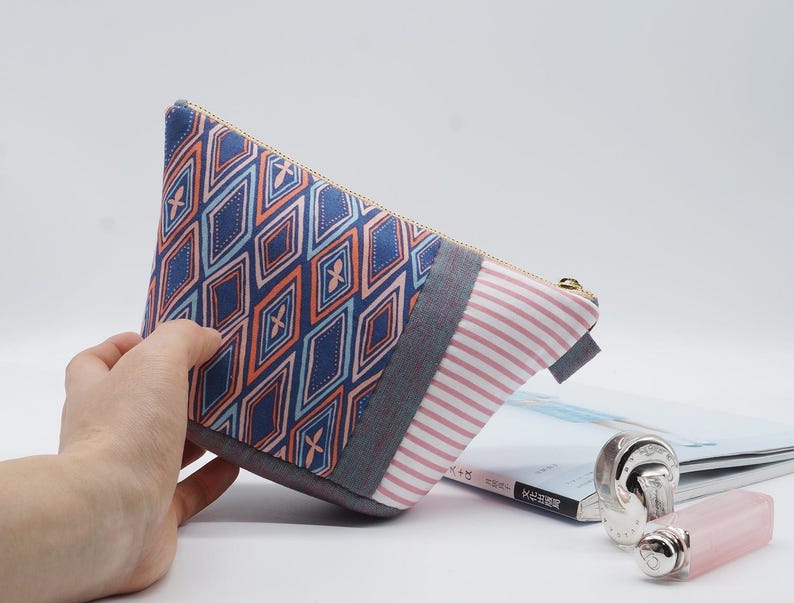 May include: A triangular cosmetic bag with a geometric pattern in blue, orange, and light blue. The bag has a striped section in pink and white, a gray accent, and a gold zipper. The bag is on a white background.
