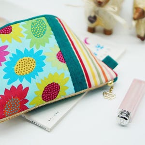 May include: A colorful cosmetic bag with a sunflower pattern. The bag features a teal green stripe and a striped pattern on the side. A gold zipper pull with a crown charm is attached. A lipstick and a ring are also visible.