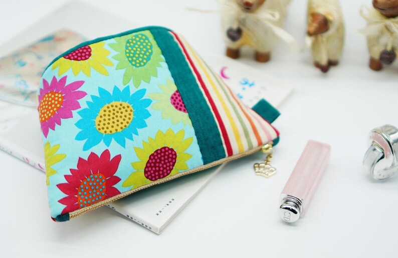 May include: A colorful cosmetic bag with a sunflower pattern. The bag features a teal green stripe and a striped pattern on the side. A gold zipper pull with a crown charm is attached. A lipstick and a ring are also visible.