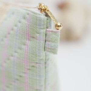 May include: Close-up of a small, quilted pouch with a gold zipper. The fabric is a light green with a subtle plaid pattern in pink and blue. The zipper pull is a small gold ball.