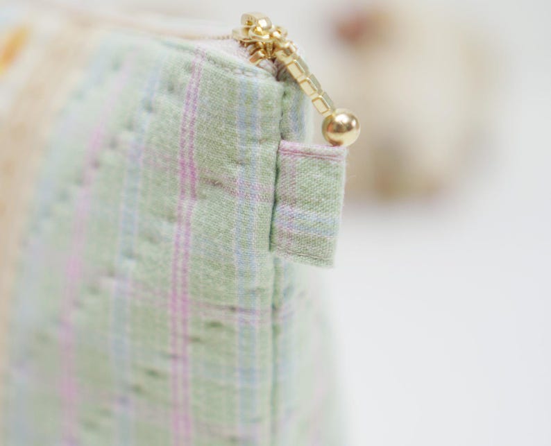 May include: Close-up of a small, quilted pouch with a gold zipper. The fabric is a light green with a subtle plaid pattern in pink and blue. The zipper pull is a small gold ball.