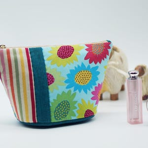 May include: A colorful cosmetic bag with a sunflower pattern and a striped side panel. The bag is teal, blue, pink, yellow, and white. A pink lipstick and a silver compact are in front of three decorative sheep.