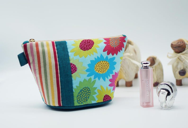 May include: A colorful cosmetic bag with a sunflower pattern and a striped side panel. The bag is teal, blue, pink, yellow, and white. A pink lipstick and a silver compact are in front of three decorative sheep.
