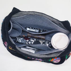 May include: A black tote bag with a colorful floral design around the rim. The bag's interior features a gray felt organizer with multiple compartments holding various items, including a wallet, phone, and a white container.