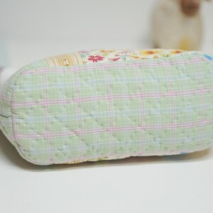 May include: A quilted fabric pouch with a light green plaid pattern on the bottom. The top of the pouch features a patchwork design with floral and patterned fabrics. The pouch is on a white surface.