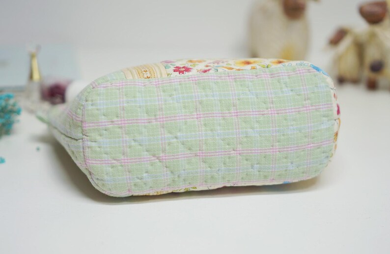 May include: A quilted fabric pouch with a light green plaid pattern on the bottom. The top of the pouch features a patchwork design with floral and patterned fabrics. The pouch is on a white surface.