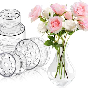 May include: Four clear plastic flower frog rings of different sizes, designed to hold flower stems in place. The rings are stacked on top of each other. A clear glass vase with a bouquet of pink and white roses is in the background.