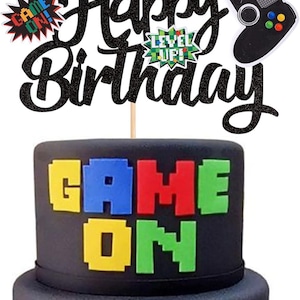 May include: A two-tiered black cake decorated with a video game theme. The top of the cake has the words "GAME ON" in colorful pixelated letters. The cake is topped with a "Happy Birthday" cake topper and a controller.