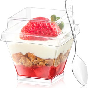 May include: A clear plastic dessert cup with a lid, filled with layers of red fruit, granola, and white cream, topped with a fresh strawberry. A clear plastic spoon rests beside the cup. The dessert is presented on a white surface.