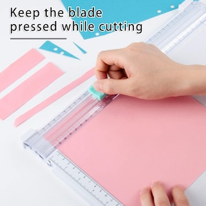 May include: A white paper trimmer with a clear ruler and a green slider. The ruler is marked in centimetres. A hand is holding the slider down while cutting a pink piece of paper.
