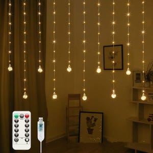 Op de afbeelding: Warmwitte LED-slingerverlichting met stervormige accenten en bolvormige lampen. Inclusief afstandsbediening en USB-connector. De lichten hangen voor een gordijn en een muur met ingelijste kunst.