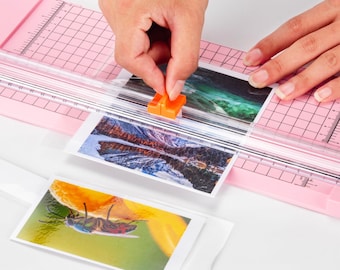 Puede incluir: Un cortador de papel rosa con una cuchilla de corte transparente y un deslizador naranja. El cortador se utiliza para cortar fotos, papel y otros materiales. Hay tres fotos en el cortador, una de una cascada, una de una playa y una de una abeja.