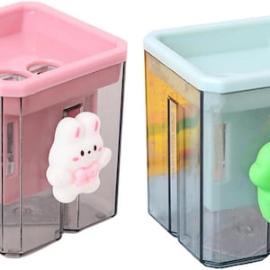 May include: Two pencil sharpeners, one pink and one pale blue, with transparent plastic bodies. The pink sharpener has a white bunny charm, and the blue one has a green dinosaur charm. Both have a square top.