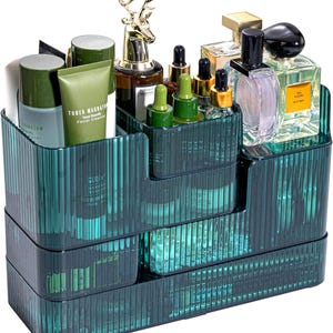 Puede incluir: Un organizador de cosméticos escalonado de color verde azulado, lleno de varios productos de belleza. El organizador tiene múltiples compartimentos y está hecho de un material translúcido acanalado. Una figura de ciervo dorado se encuentra encima.