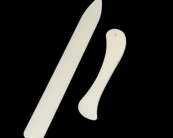 May include: Two white plastic tools for paper crafts. One is a long, thin blade with a rounded tip. The other is a shorter, curved blade with a hole in the handle.