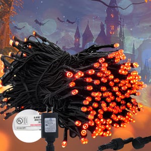 Puede incluir: Cadena de luces LED naranja con cableado negro, ideal para Halloween o decoraciones festivas. Las luces están agrupadas, con un adaptador de corriente y conector visibles. El fondo presenta una escena de castillo espeluznante.