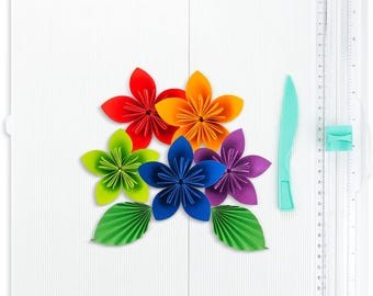 Puede incluir: Una estera de corte autocicatrizante blanca con una regla en el lateral. Hay seis flores de papel de colores y dos hojas de papel verde en la estera. Un cuchillo de manualidades de plástico turquesa se encuentra en el lado derecho de la estera.