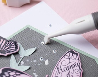 Puede incluir: Una herramienta blanca con un mango gris se utiliza para presionar lentejuelas plateadas sobre un fondo de cartulina gris. La cartulina está decorada con una mariposa rosa y negra, hojas verdes y salpicaduras de pintura blanca. La cartulina está rodeada por un borde verde. La cartulina está sobre una superficie rosa. El texto "Happy" es visible en la cartulina.