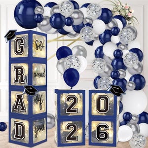 May include: A graduation party decoration set featuring navy blue boxes spelling out "GRAD" and "2026", with a "so proud of you" message. The set includes navy, silver, and white balloons, and graduation cap accents.