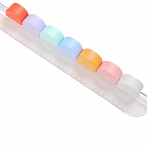 Puede incluir: Una regla de plástico transparente con formas de semicírculo de colores arcoíris unidas a la parte superior. La regla tiene marcas en pulgadas.