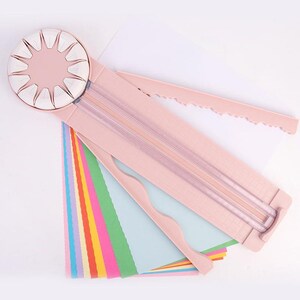 Pode incluir: Cortador de papel rosa com uma lâmina rotativa branca e uma régua. O cortador tem uma borda ondulada e uma borda serrilhada. Há várias folhas de papel colorido sob o cortador.