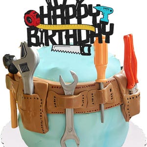 Peut inclure: Un gâteau bleu clair décoré d'une ceinture à outils marron et de divers outils. Le gâteau est surmonté d'un cake topper noir pailleté "Happy Birthday" avec des illustrations d'outils. Une clé anglaise argentée et un tournevis orange sont visibles.