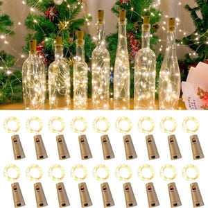 May include: Clear glass bottles with cork stoppers, each filled with warm white fairy lights. Below, several sets of cork-shaped battery packs and light strings are arranged. The background features a Christmas tree with more lights.