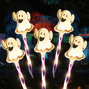 Könnte beinhalten: Fünf Halloween-Leuchtstäbe. Jeder Stab hat ein lächelndes Geisterdesign mit braunem Rand und einem lila-weiß gestreiften Griff. Im Hintergrund ist ein Friedhof mit den Worten "BE RE" zu sehen.